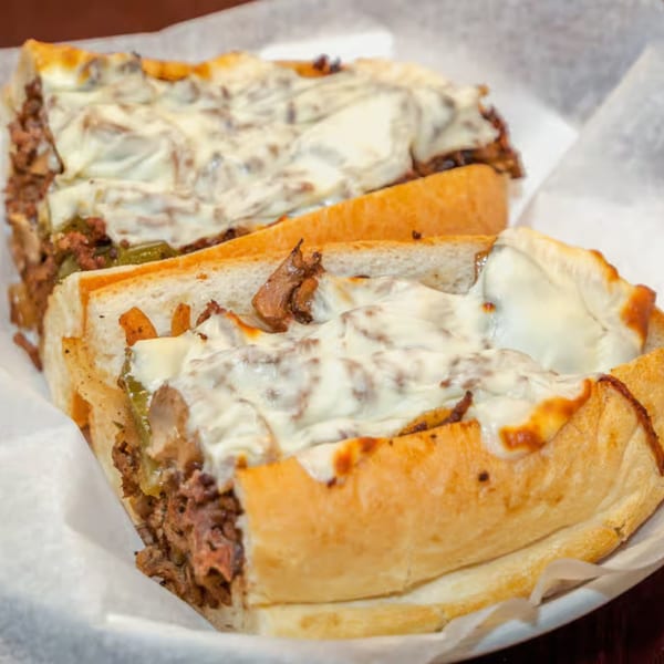 Two halves of a cheesy steak and pepper sub sandwich on a white paper-lined tray.