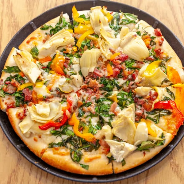 Antigoni's Pizza with artichokes, bell peppers, bacon, and spinach on a black pan.